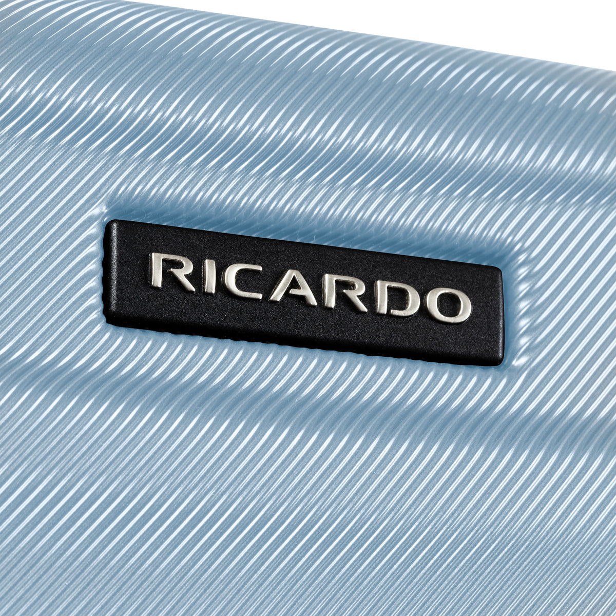A close-up of a light blue textured hard case with a black rectangular brand plaque reading RICARDO in silver lettering.