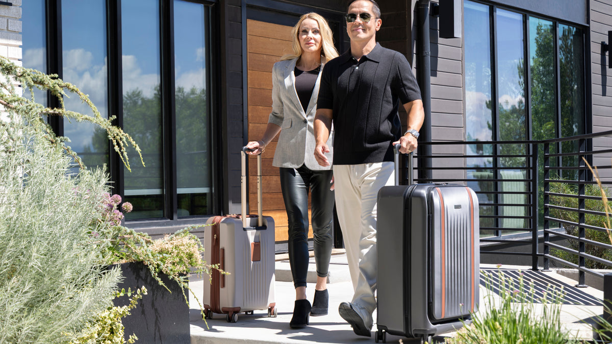 A man and a woman walk out of a modern building, each pulling a hard-shell rolling suitcase. The man wears a black polo shirt and light pants, and the woman wears a light blazer and dark pants.