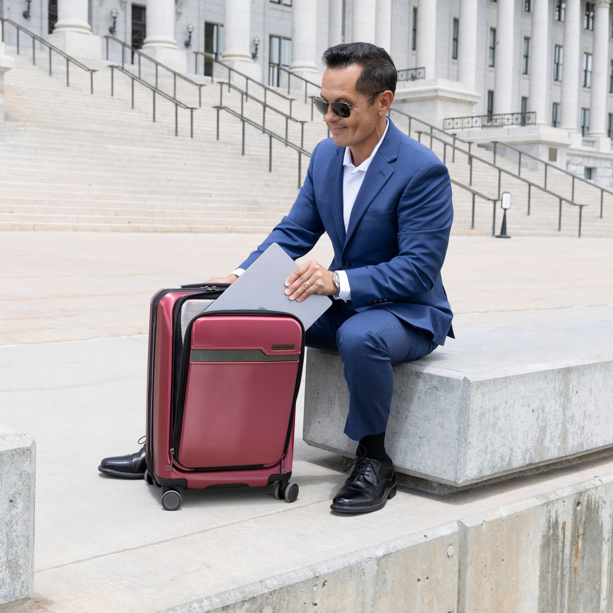 Man in a navy suit sitting on stone steps, placing a laptop into the front compartment of a burgundy Ricardo hardside carry-on suitcase, with a capitol building behind him.